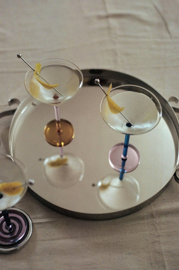Three colorful stemmed martini glasses with lemon twists on a round mirrored tray