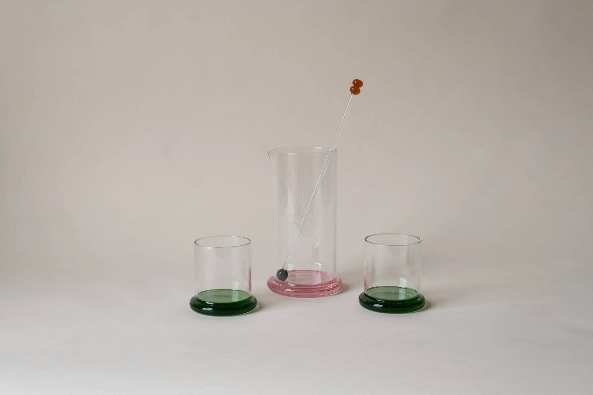 Modern glass pitcher with pink base, glass stir stick and two green-base tumblers on neutral background
