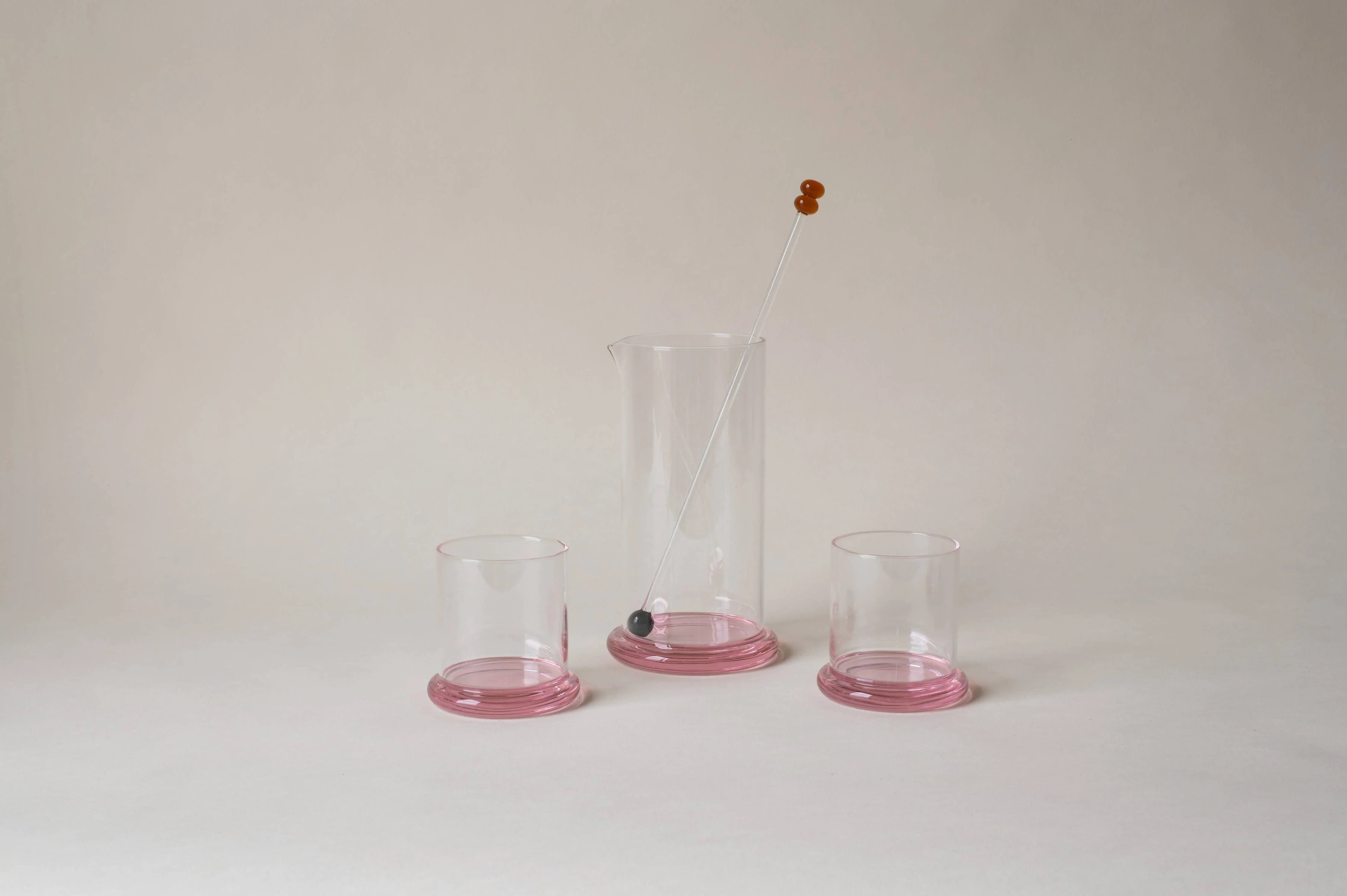 Clear glass pitcher and two tumblers with pink bases, glass stirrer on neutral background.