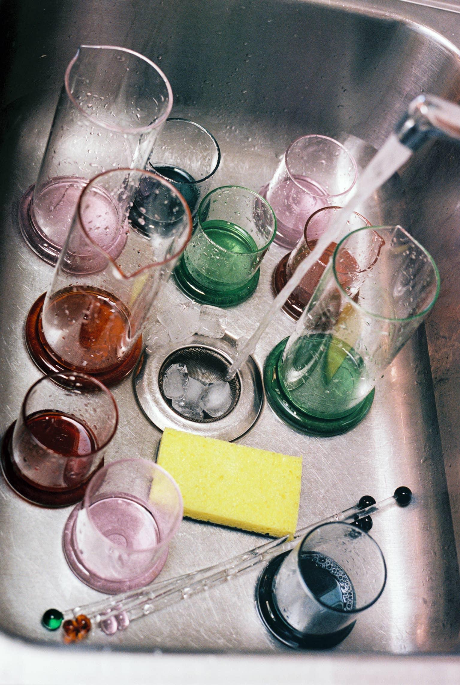 Colorful glass cups, pipettes, a yellow sponge, and running water in a stainless steel sink
