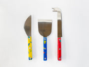 Three cheese knives with speckled yellow, blue, and red handles laid out on white