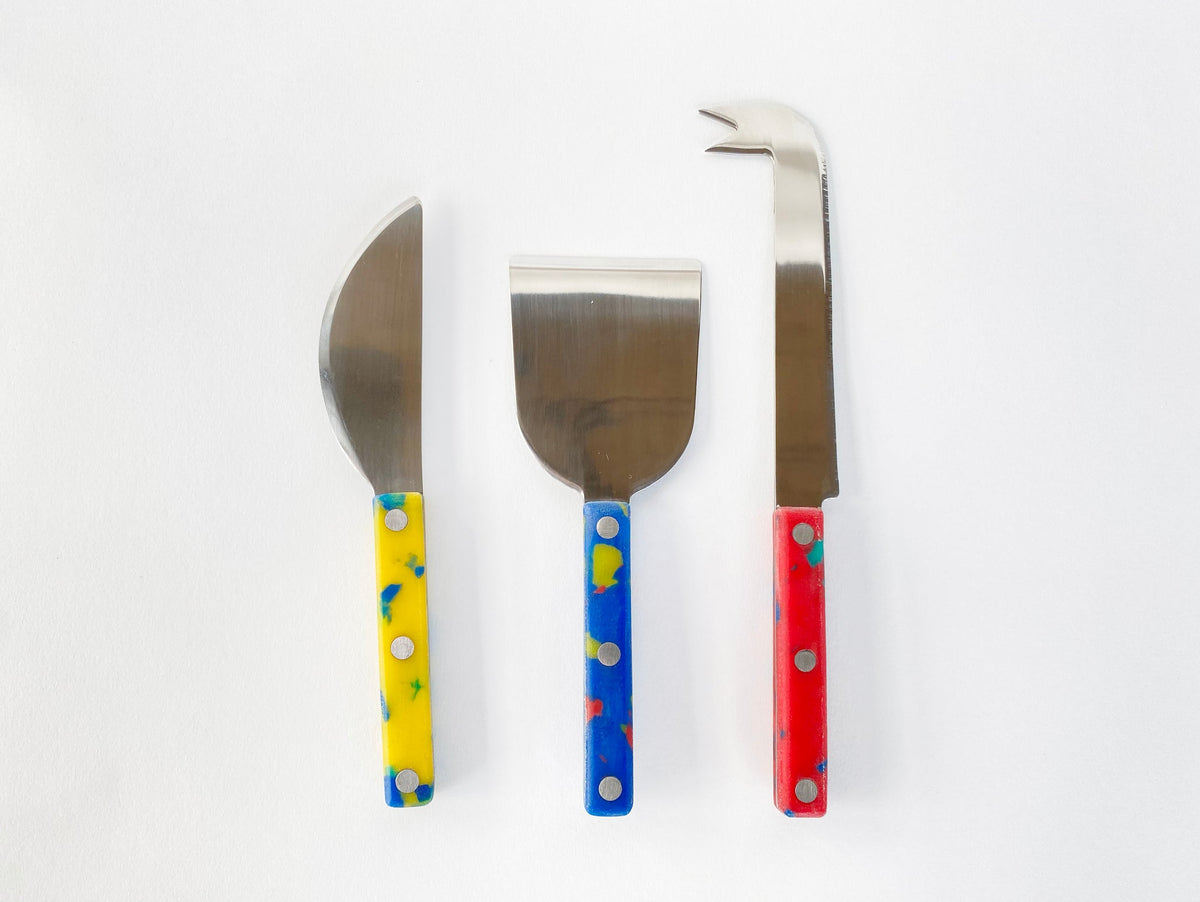 Three cheese knives with speckled yellow, blue, and red handles laid out on white