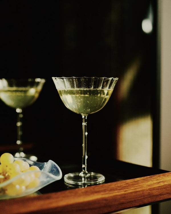 Elegant scalloped champagne coupe glass with bubbles beside grapes on wooden tray