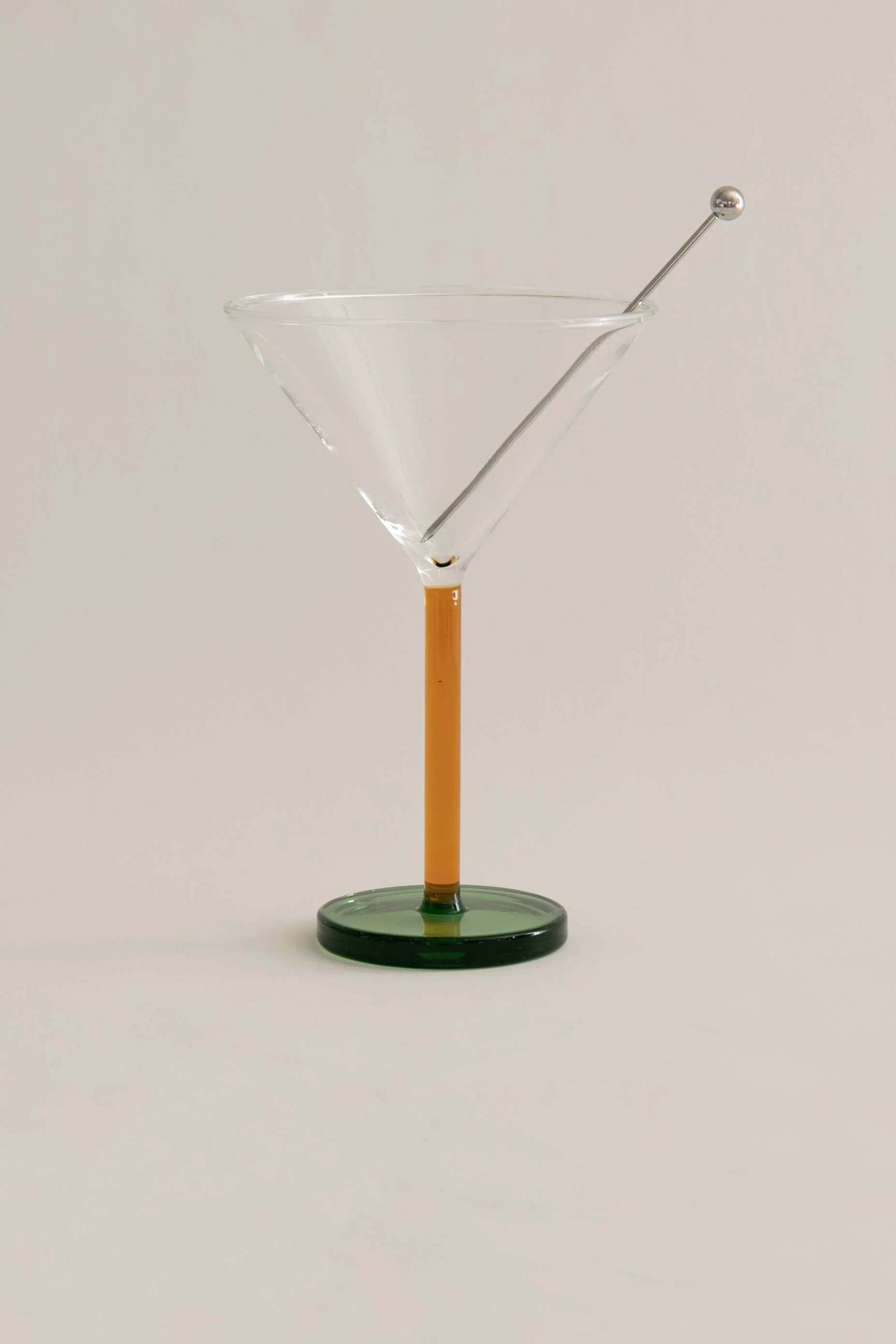 Modern martini glass with orange stem, green base, and metal cocktail pick on white background