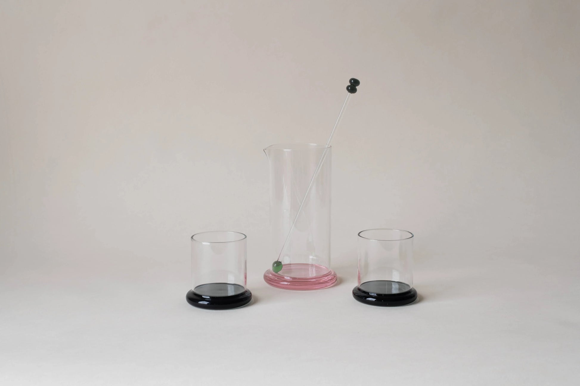 Modern glass cocktail set with pink pitcher, black-based glasses, and mixing stick on white background