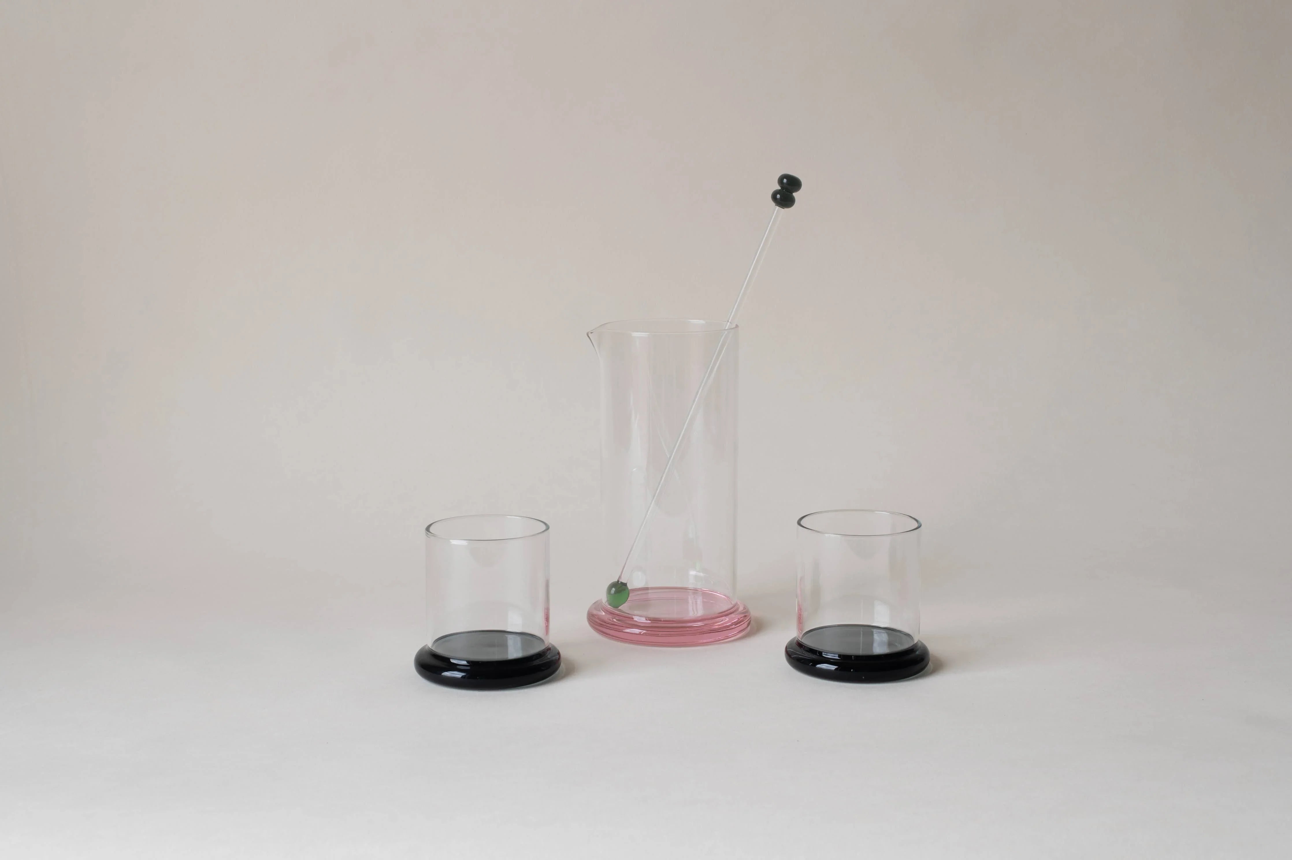 Modern glass cocktail set with pink pitcher, black-based glasses, and mixing stick on white background