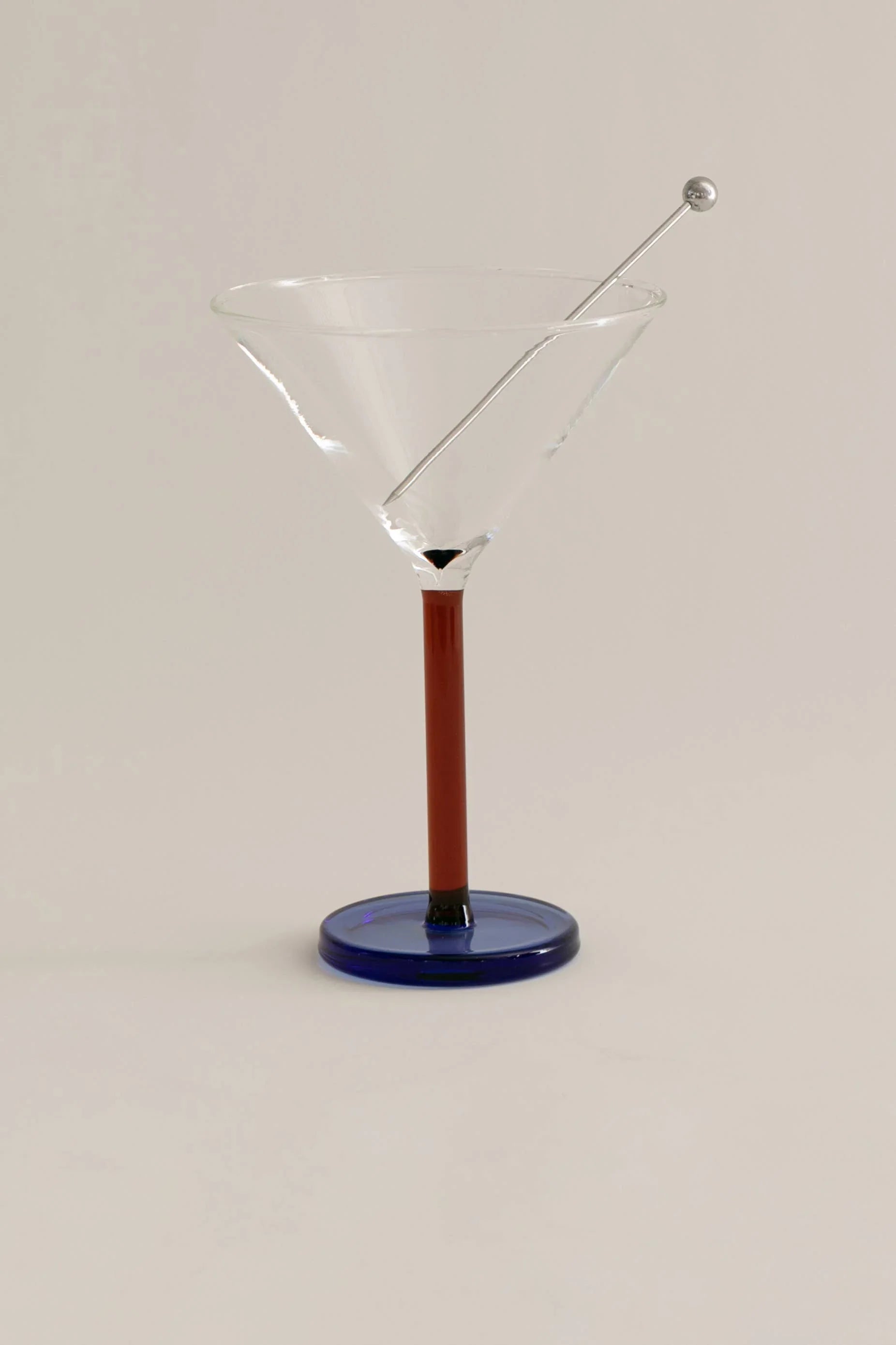 Clear martini glass with a metal stirrer, brown stem, and blue base on white background